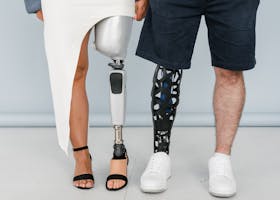Two individuals with stylish clothing and prosthetic legs standing together on a minimalist background.