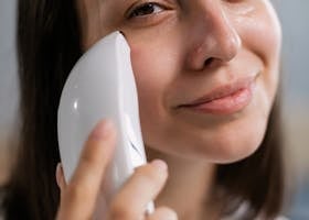 A close-up of a smiling woman using a facial skincare device indoors for healthy skin.